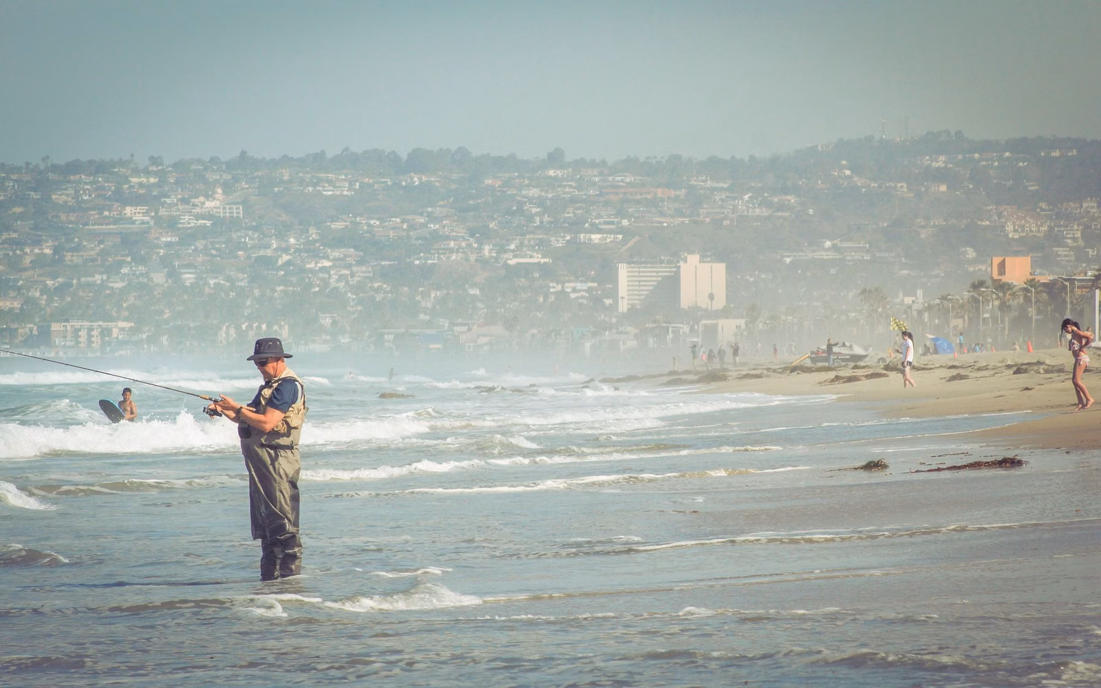 An angler in waders surf fishing in shallow water off a Southern California beach
