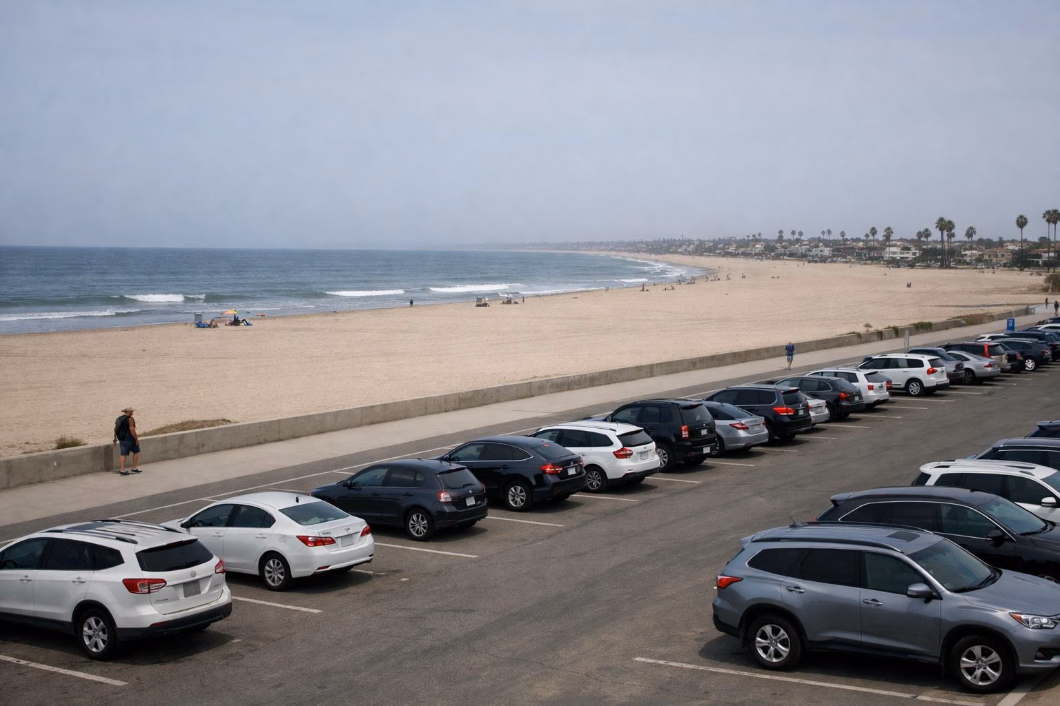 A paved beach parking lot directly behind a wide sandy beach, with the ocean beyond